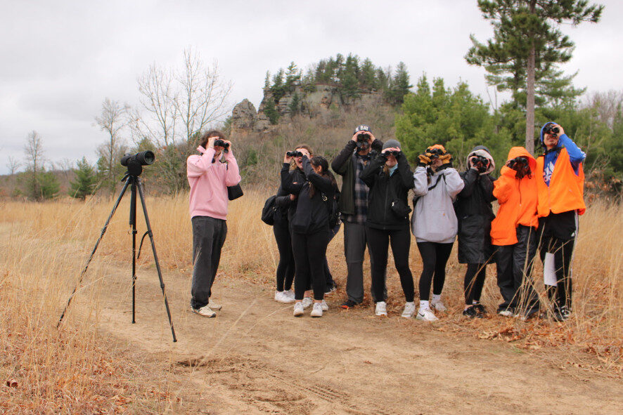 students looking through binoculars and standing on a sandy and grassy trail under cloudy skies