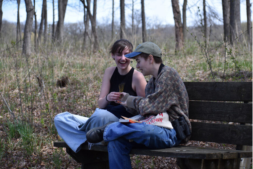 two people sitting on a bench in the woods looking at bird feathers