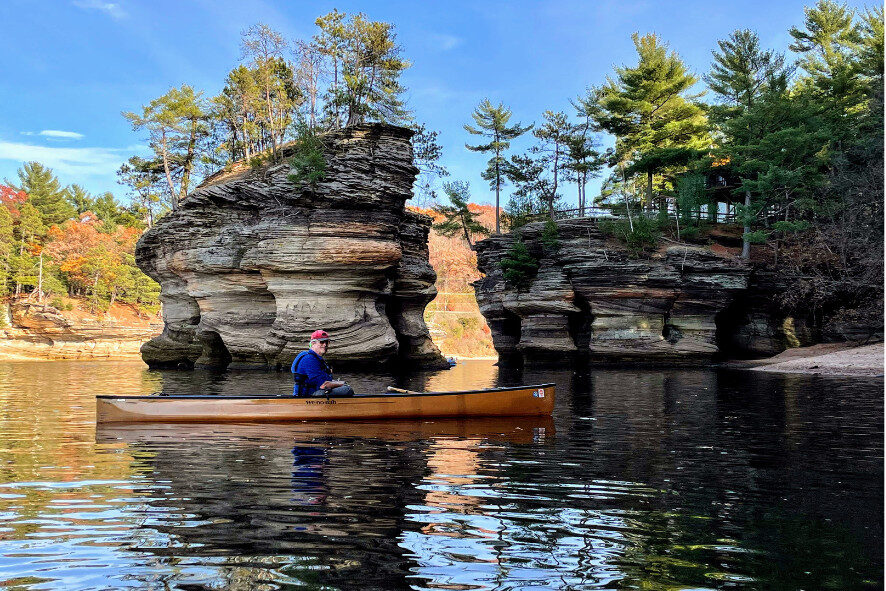 Person canoeing in clear water around large rock formations and pine trees